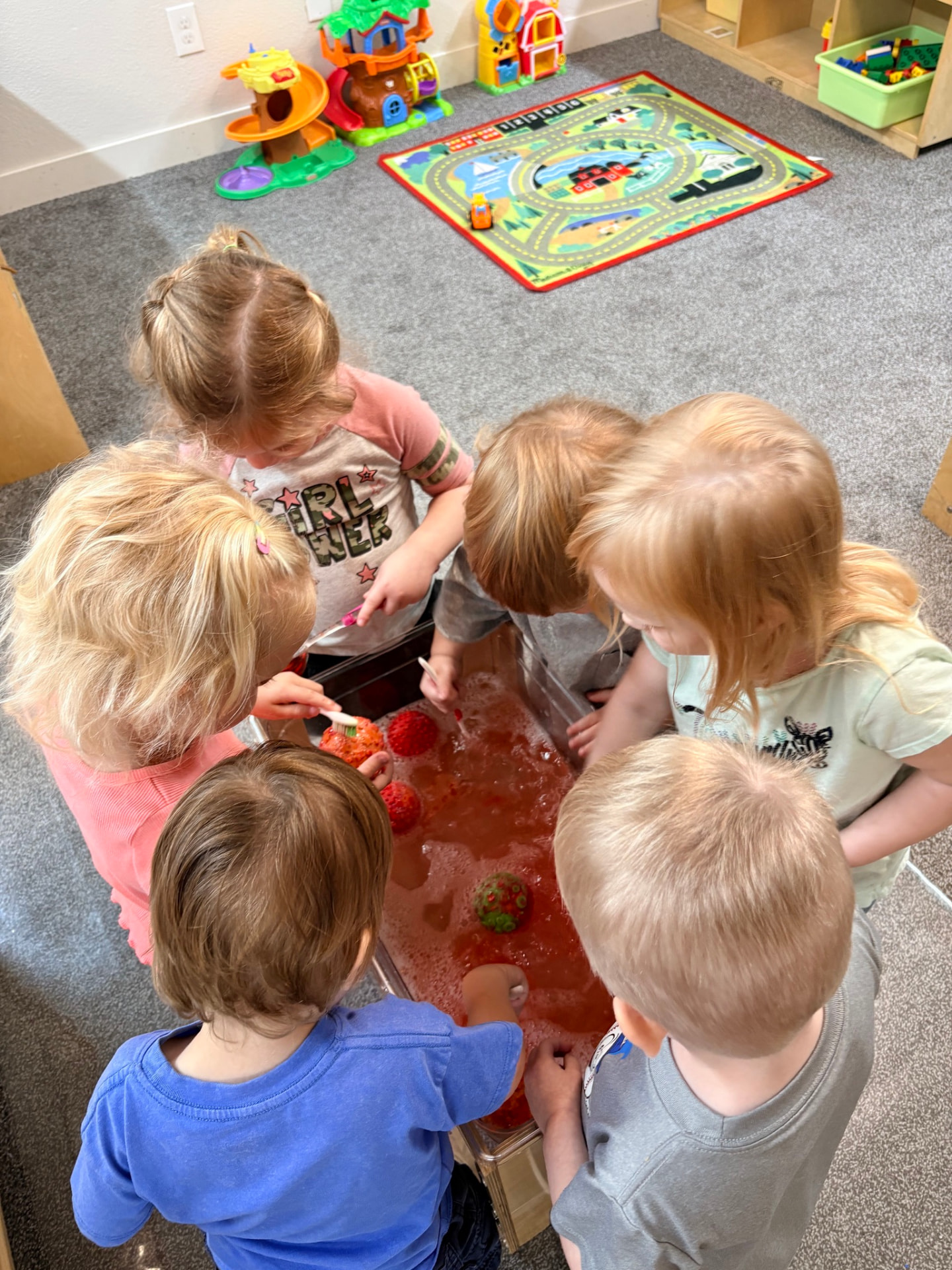 Six toddlers gathered around a bubbly red sensory water table exploring together in the Chickadees room at The Nest