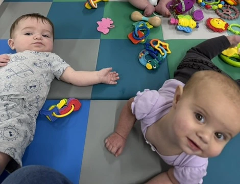 Two infants playing on a colorful foam play mat with bright toys in the Owlets infant room at The Nest